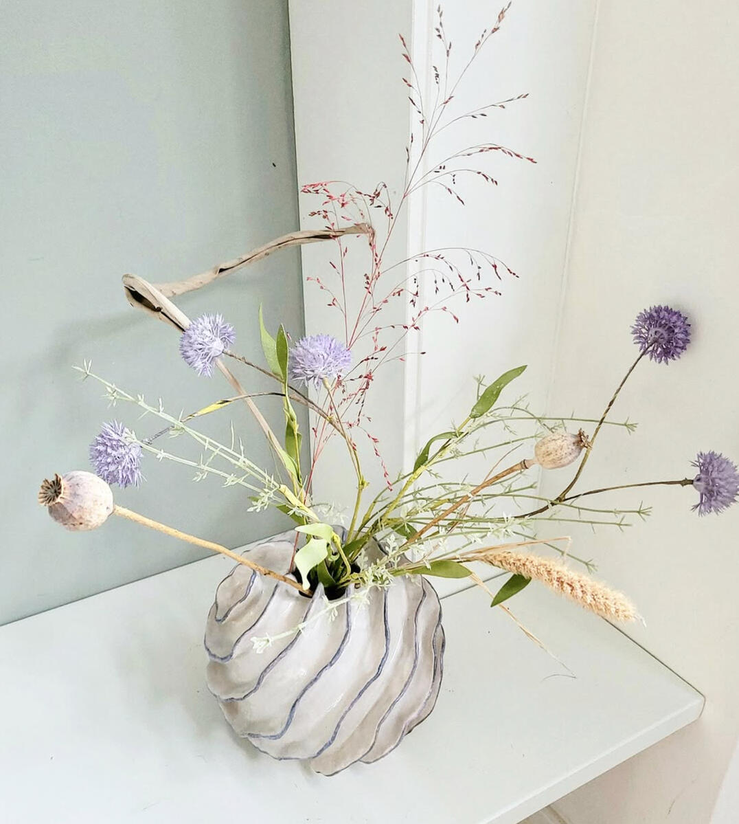 Wavy relief ceramic vase — handbuilt floral vessel by Natty Ceramics atelier in Utrecht Handbuilt ceramic vase with ribbed wavy relief, evoking flowing water or sand dunes, holding dried flowers with poppy pods and purple alliums